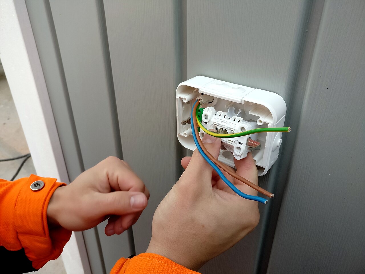 Les mains d’un électricien en tenue orange branchent les fils d’une prise murale.
